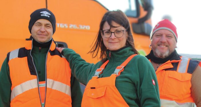 Zwei Müllwerker und eine Müllwerkerin stehen vor einem Müllwagen.