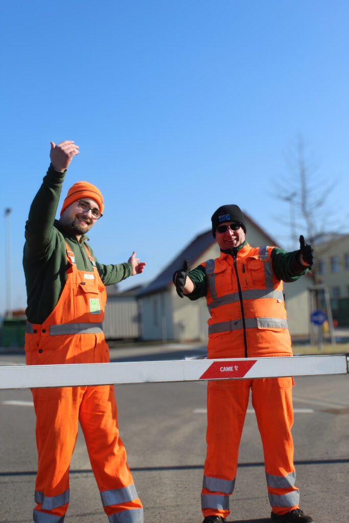 Zwei Wertstoffhofmitarbeiter stehen einladend an der Schranke zum Hof.