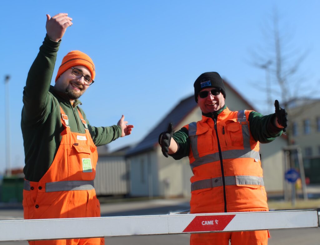 Zwei Wertstoffhofmitarbeiter stehen einladend an der Schranke zum Hof.