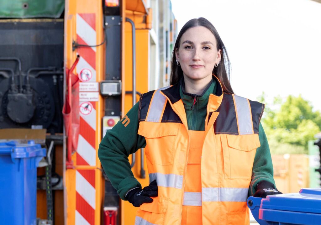 Müllwerkerin steht neben dem Müllauto mit einer blauen Tonne