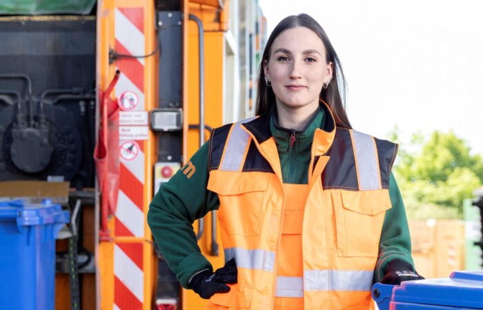 Müllwerkerin steht hinter einem Müllfahrzeug und schaut freundlich