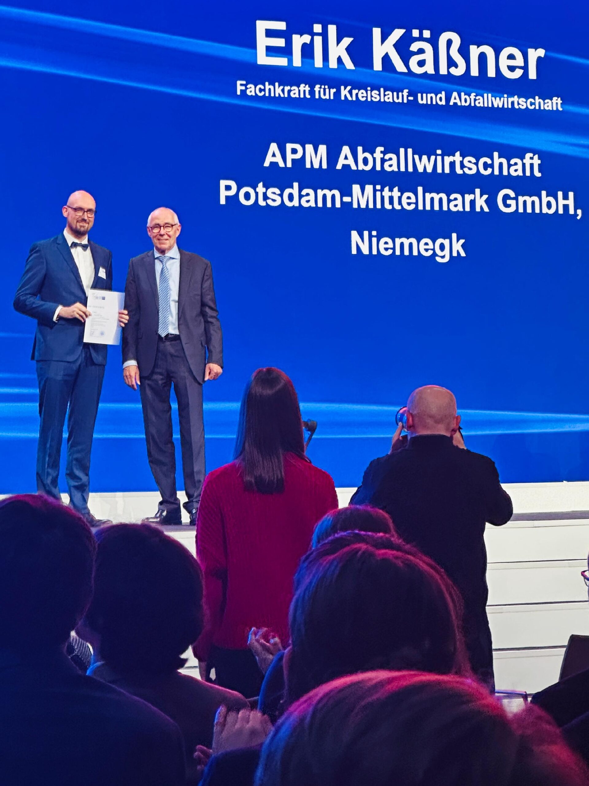 Junger Mann steht auf einer Bühne und hat seine Urkunde erhalten. 