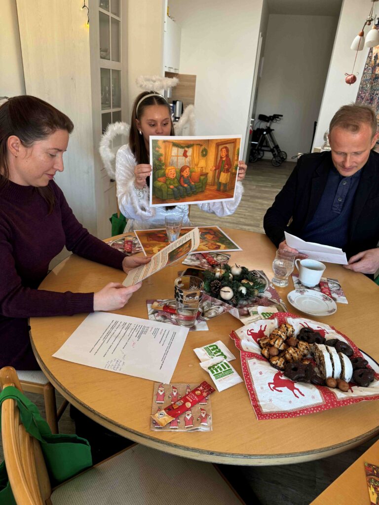 Duo liest Senioren eine Weihnachtsgeschichte vor während ein verkleideter Engel die Bilder zur Geschichte hoch hielt.